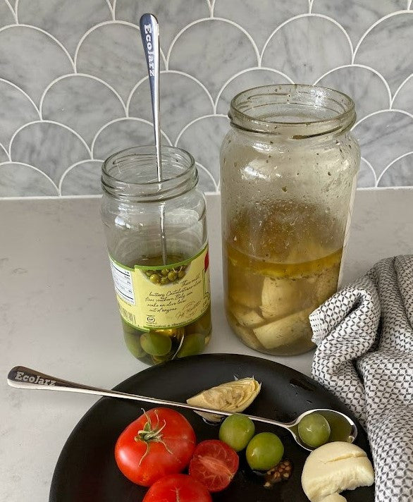 ecojarz skinny spoon reaches the bottom of a jar of olives, on a kitchen counter next to a plate of charcuterie