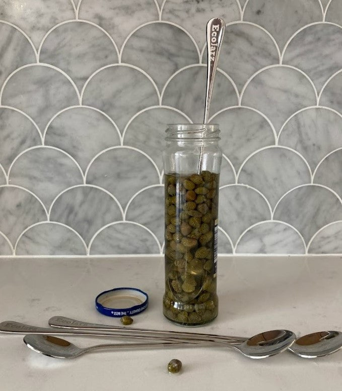 ecoJarz stainless steel skinny spoon resting in a tall, thin jar of capers, it reaches the bottom of the jar