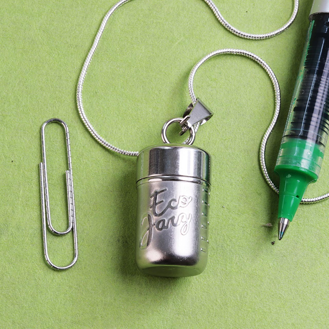 Stainless Steel Mason Jar Locket Necklace laying next to a paper clip and a pen for size comparison.