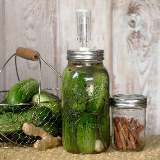 A large mason jar filled with pickles with an airlock and fermenter lid next to a basket of cucumbers.